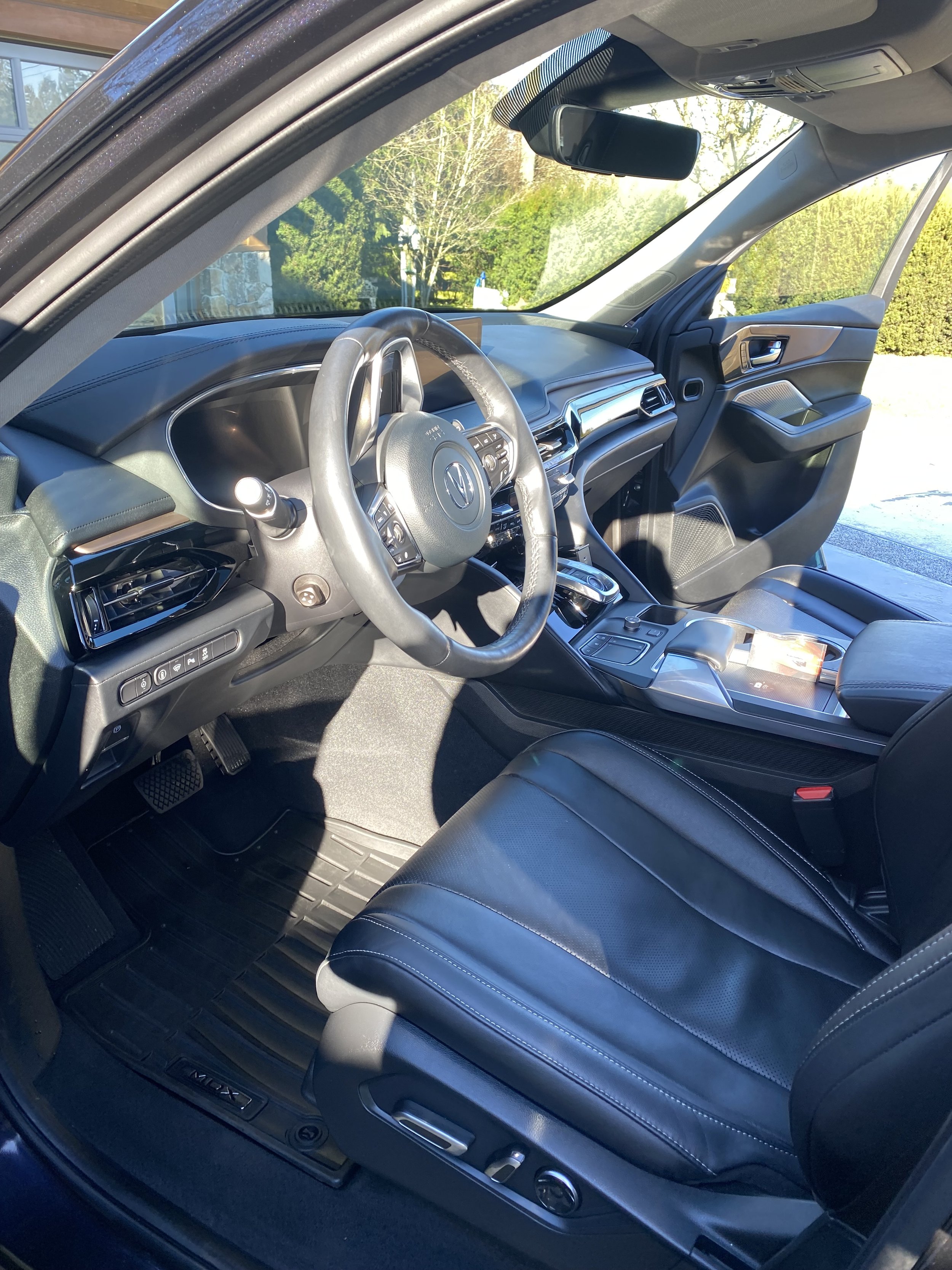 Interior of a modern vehicle showing the driver's seat, steering wheel with controls, dashboard, center console with gear shift, and front passenger seat. Outside view through the windshield includes trees and a building.