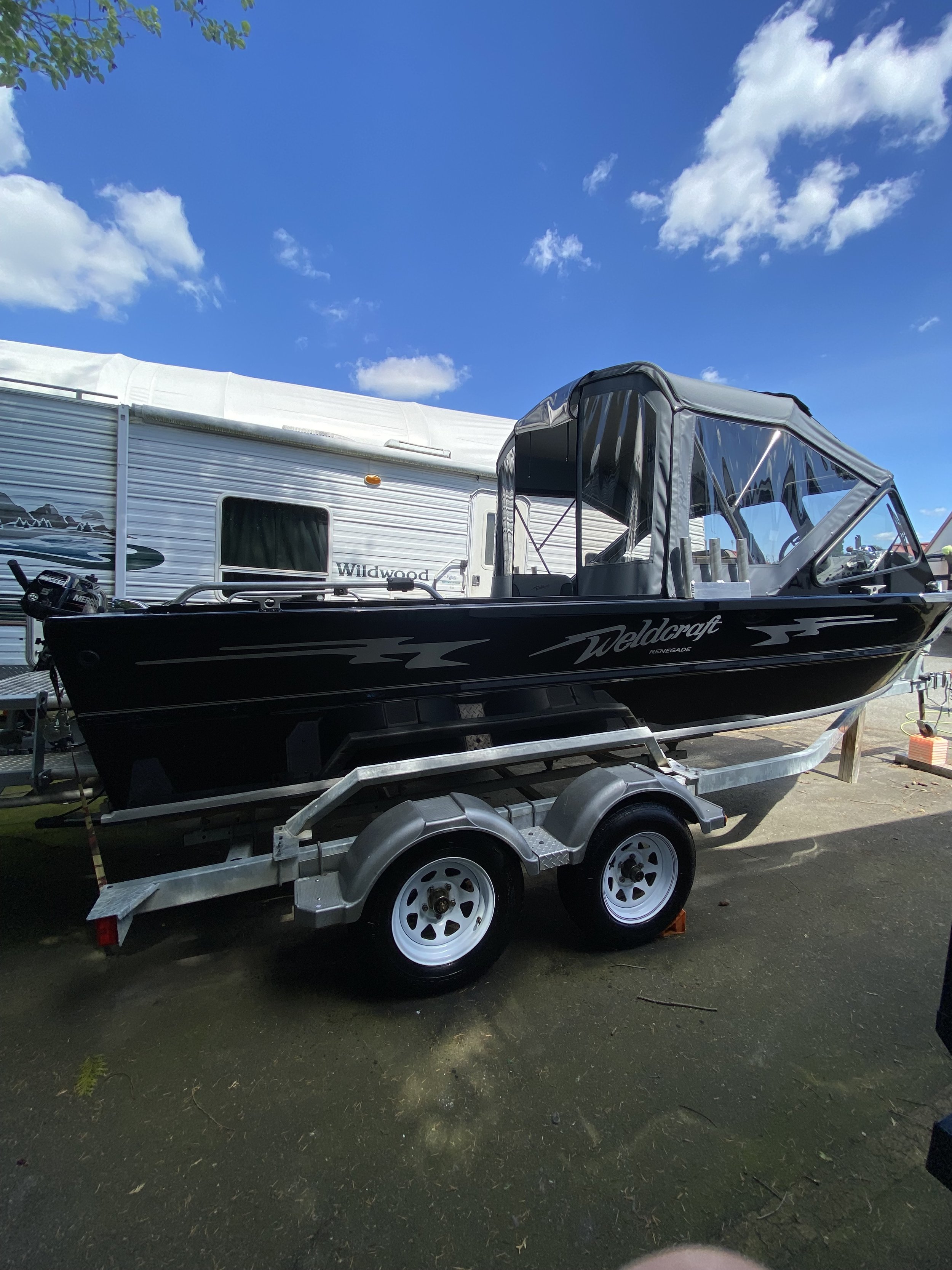 Black Weldcraft Renegade boat on a double-axle trailer next to a white Wildwood RV under a blue sky with scattered clouds.