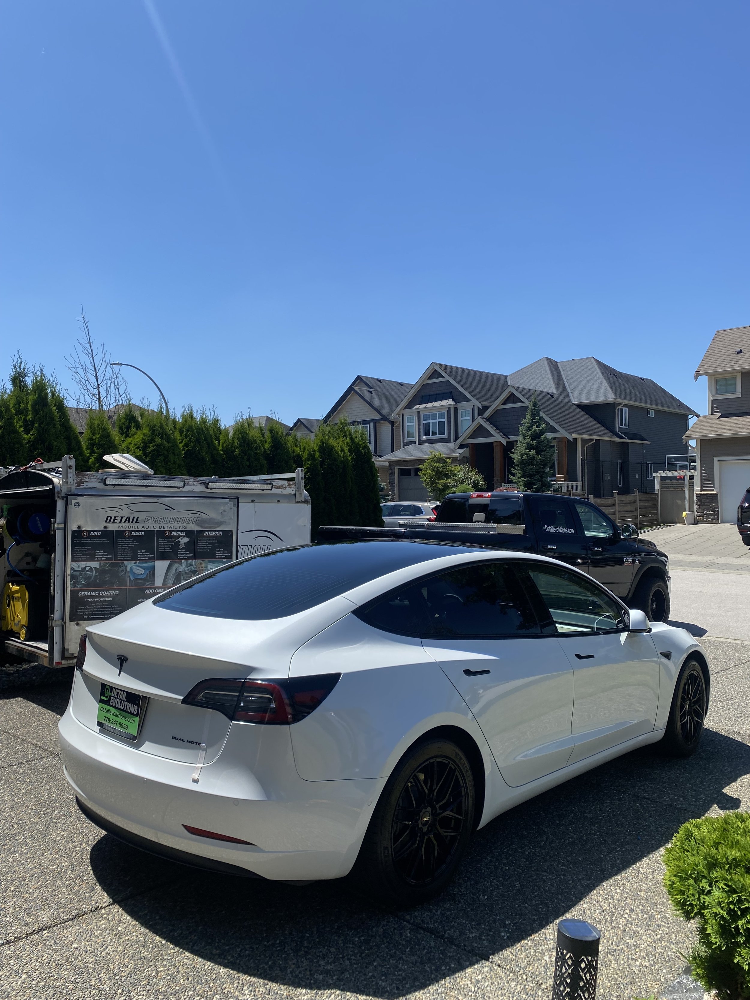 White Tesla car parked in driveway with a black SUV behind it, residential neighborhood, blue sky, and a detailing truck nearby.