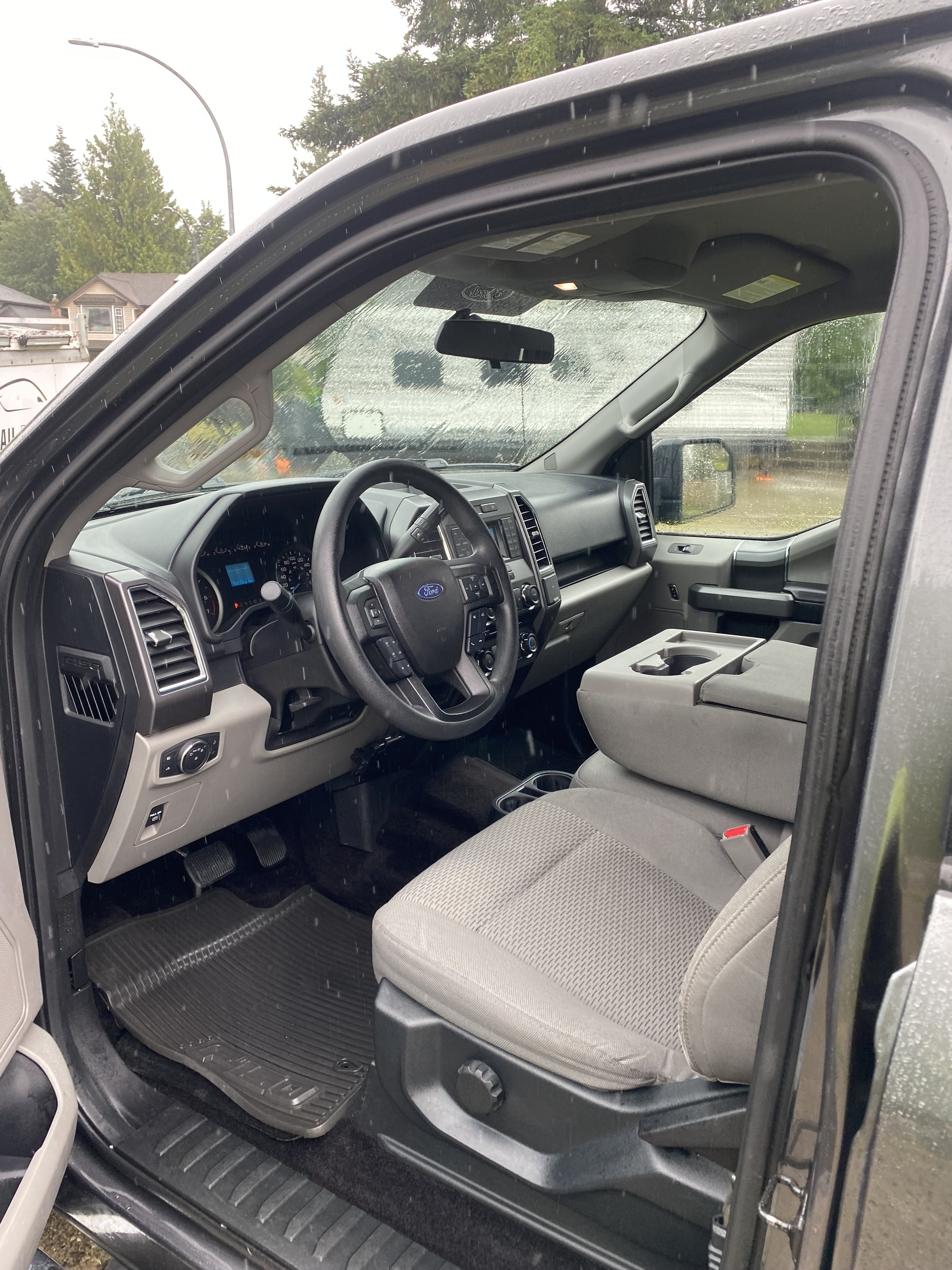 Inside view of a Ford truck cabin showing gray fabric seats, steering wheel, dashboard, and center console, with rain on the windshield.