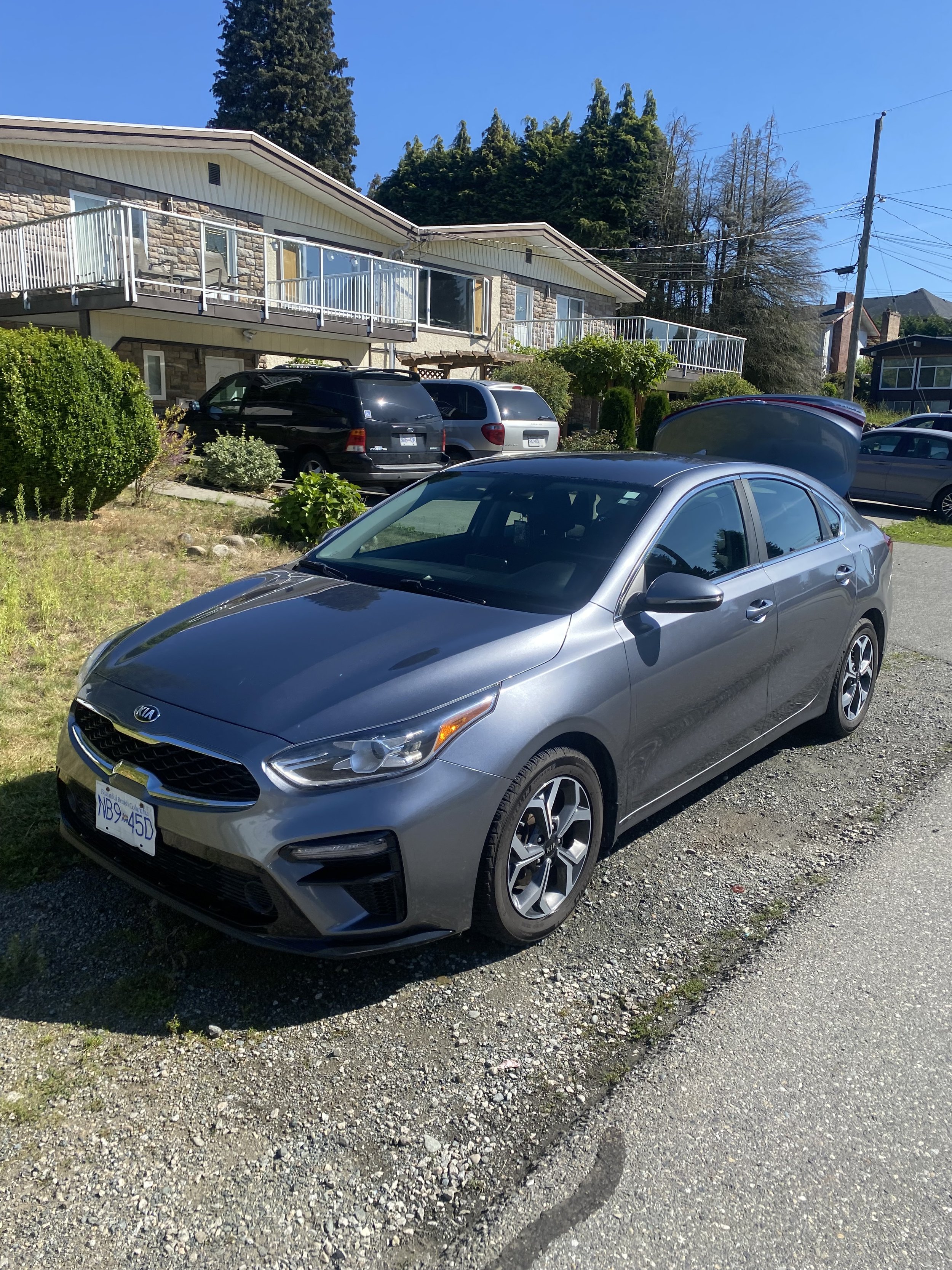 A silver Kia sedan parked on a gravel area in front of a multi-level residential building with a stone and siding exterior. In the background, several other vehicles are parked, and there are lush green trees and bushes around the building. The sky is clear and blue.