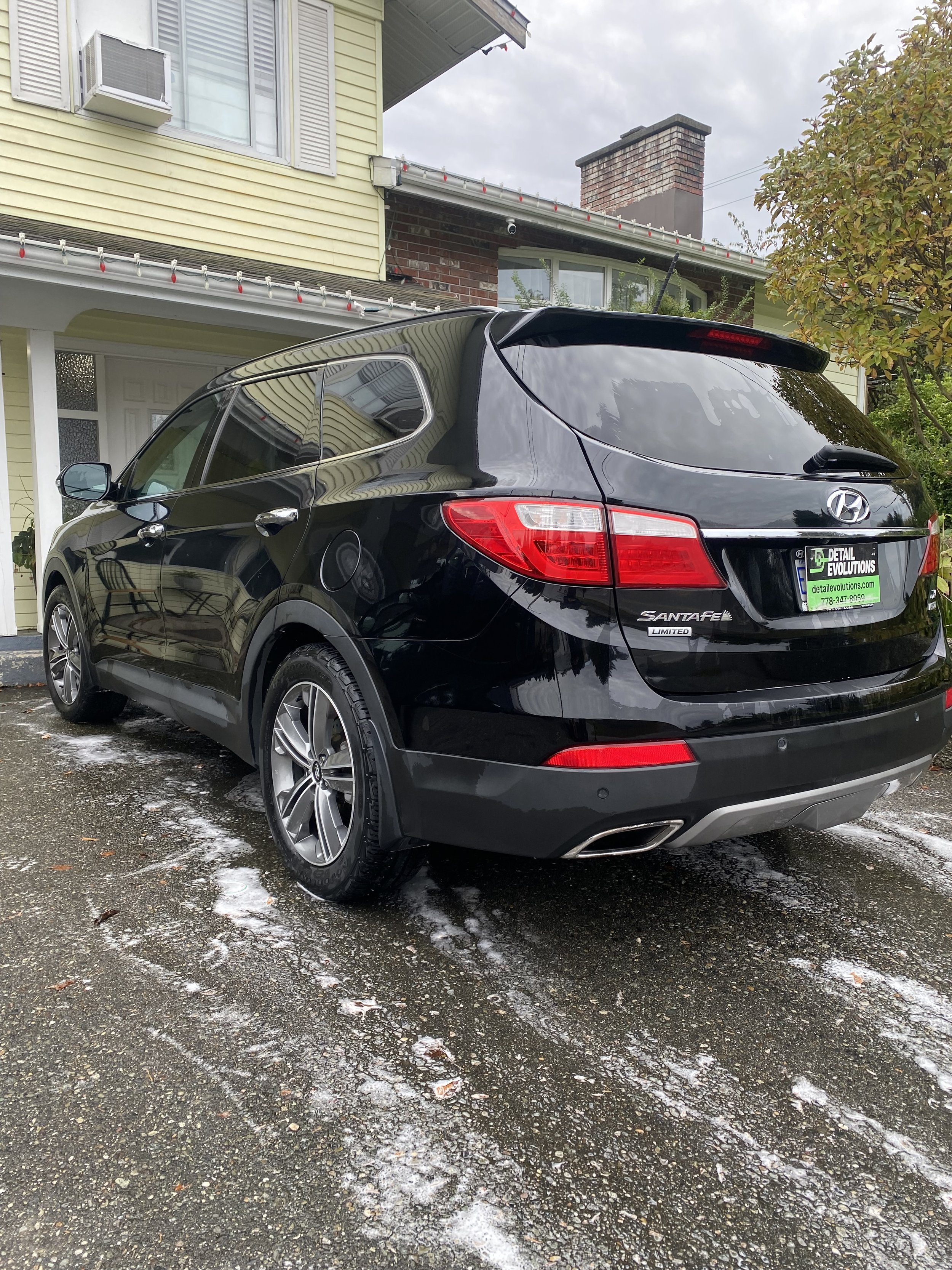 Black Hyundai Santa Fe Limited parked on wet driveway with soap suds, in front of a house with beige siding and white window shutters, overcast sky.